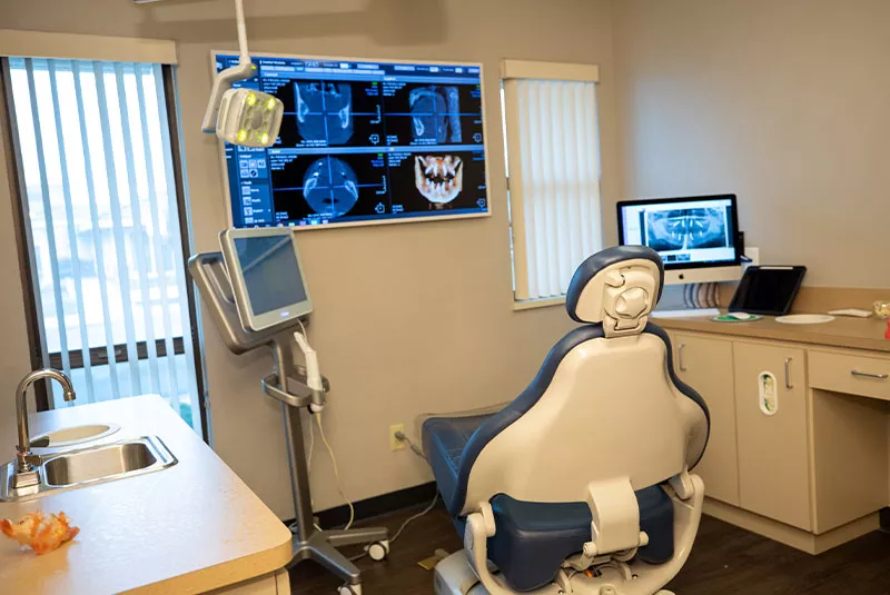 A well-equipped dental examination room with an empty patient chair, dental light, and a sink. On the wall, a large screen displays detailed dental CT scans, while a secondary monitor shows additional X-ray images. The room is designed for comfort and efficiency, with natural light coming from a window with blinds.
