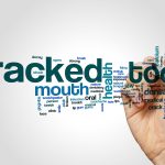 A word cloud focused on the terms "cracked tooth," with related words in various sizes, such as "mouth," "health," "dental," "jaw," and "hygiene." The text appears in shades of blue and black, with larger words indicating higher relevance. A hand on the right side of the image is holding a marker as if drawing or writing the words. The background is a plain white, keeping the focus on the text.