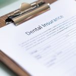 A person filling out a dental insurance form on a clipboard, with a pen in hand. The form is titled "Dental Insurance" at the top. A computer keyboard and a small model of a tooth are visible in the background on a glass desk, suggesting a dental or office setting.