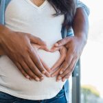 Close-up of a pregnant woman's belly with her hands and her partner's hands gently placed over it. Their fingers form a heart shape, symbolizing love and anticipation for the baby. The woman wears a white top and a blue cardigan, while the partner embraces her from behind. The background is softly blurred with natural light coming through a window.