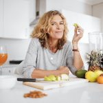 Smiling woman with curly blonde hair in a light grey top leans on a kitchen counter, holding a slice of green apple. In front of her is a cutting board with sliced apples, a knife, and assorted fresh fruits including a pineapple, mangoes, bananas, lemons, oranges, and a pomegranate. A blender, a bowl with a spoon, a lit candle, a wine glass filled with orange juice, and a small pile of almonds are also visible on the counter in a bright, modern kitchen.