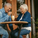 Elderly couple sitting at an outdoor café table, smiling and laughing while sharing a milkshake with two straws. Both are wearing denim outfits and appear to be enjoying each other’s company. The café has wooden walls and large windows, with a small potted plant nearby.