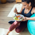 Woman in athletic wear sitting on the floor and leaning against a turquoise exercise ball, smiling while eating a colorful bowl of healthy food that includes vegetables, rice, and protein. She is wearing wireless earbuds and appears relaxed post-workout in a bright indoor space with wooden flooring.