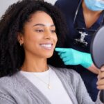 Smiling woman seated in a dental chair holds a hand mirror while a dental professional in scrubs and gloves points to her jaw during a consultation in a bright, modern dental office.